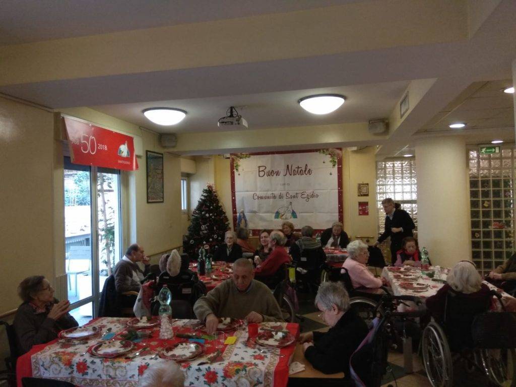 Molti anziani in sedia rotelle siedono intorno a tavoli apparecchiati per il Natale, organizzato dalla Comunità, di Sant'Egidio.