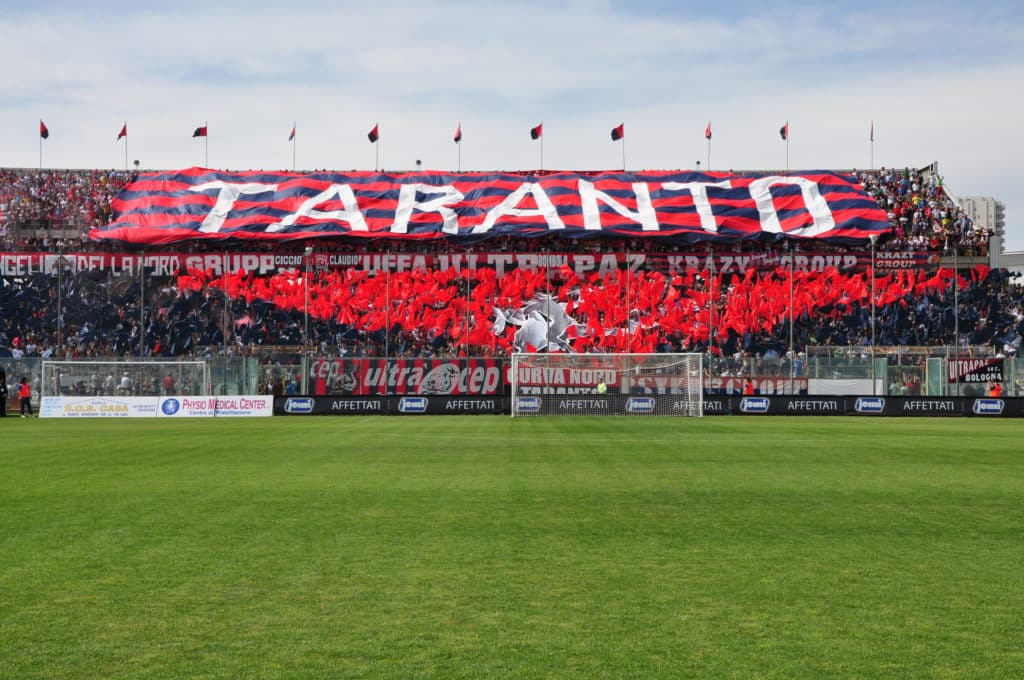 In foto la Curva Nord del Taranto. Nell'anello superiore si staglia un grande drappo a righe orizzontali rossoblu sul quale vi è scritto con grandi caratteri bianchi "Taranto". Nella parte inferiore vi è invece un gioco di bandiere rosse e blu.